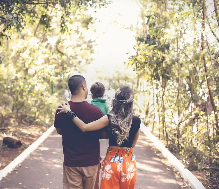 A family - man, child, woman - walks down a path lined with trees with their backs facing the camera